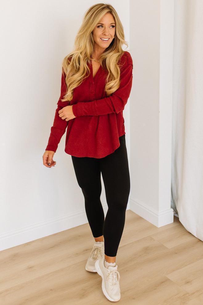 Woman wearing a red long-sleeve top and black leggings standing against a white wall.