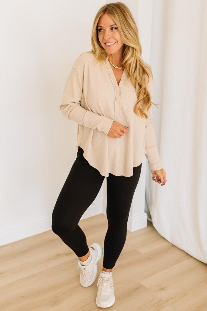 Woman wearing a beige top and black leggings standing against a white curtain.