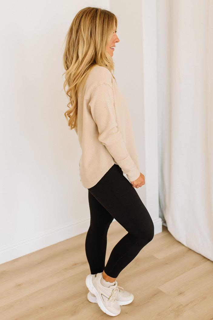 Woman wearing a beige top and black leggings standing against a white wall.