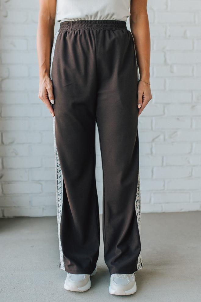 Person wearing brown pants with lace trim against a white brick wall.