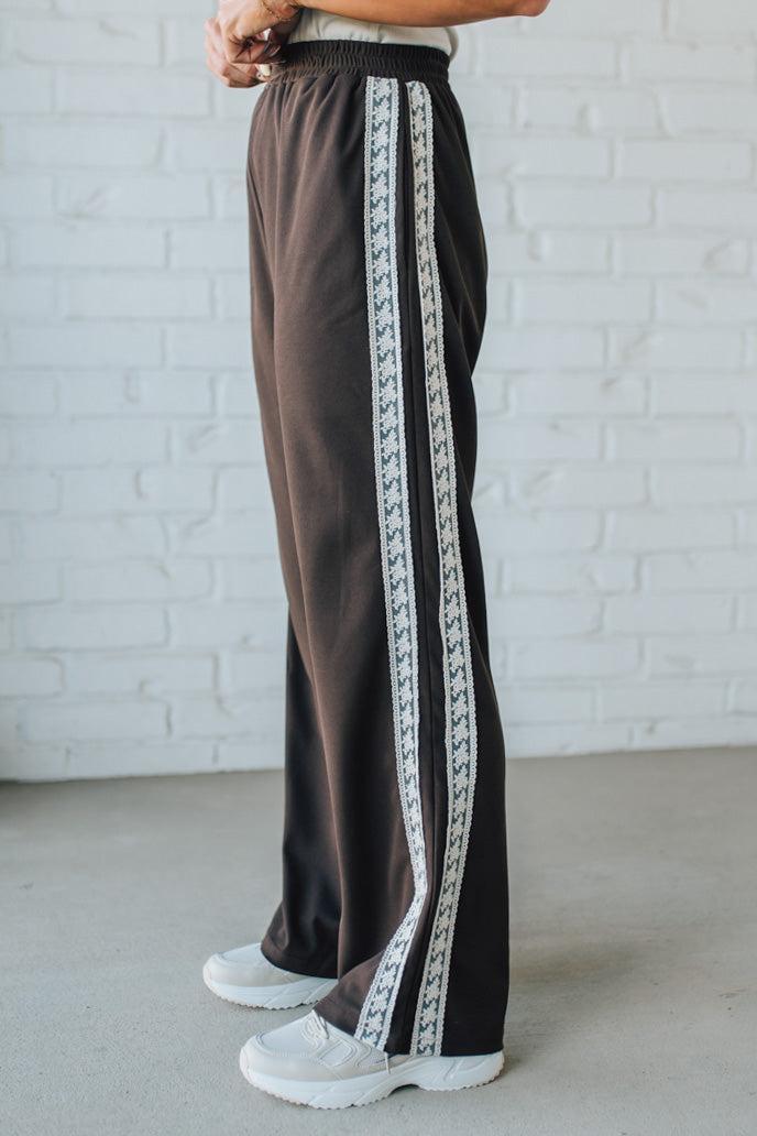 Brown pants with white lace trim worn by a person against a white brick wall.