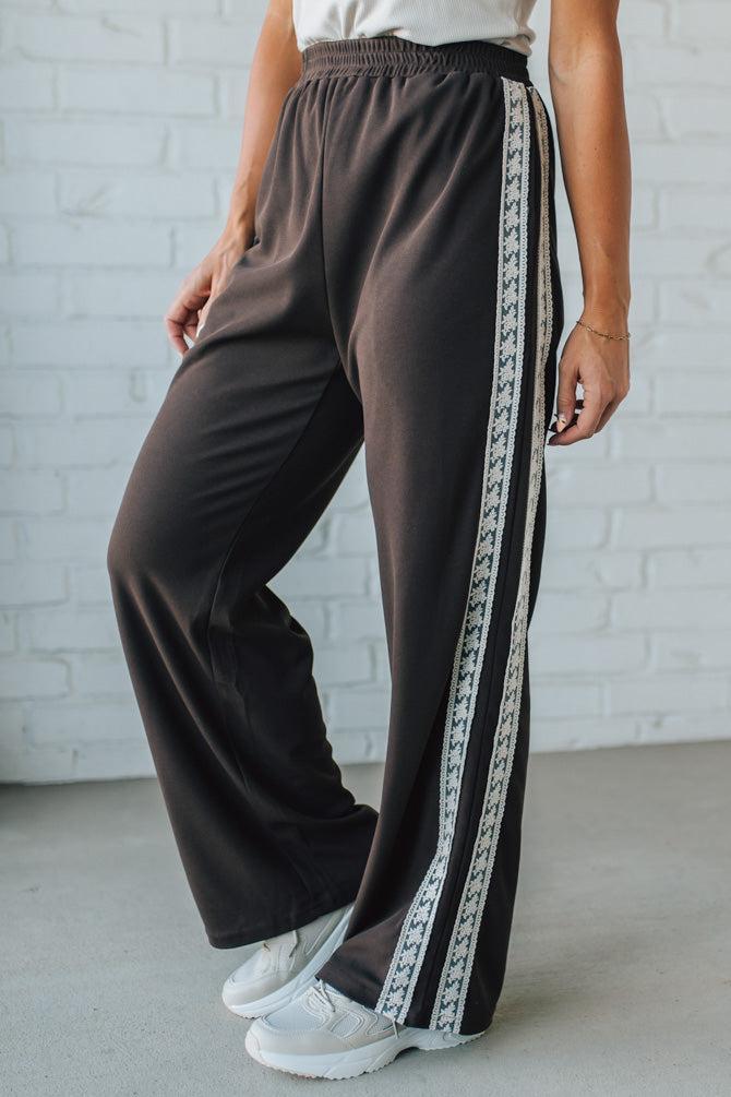 Brown wide-leg pants with a decorative stripe worn by a person against a white brick wall.