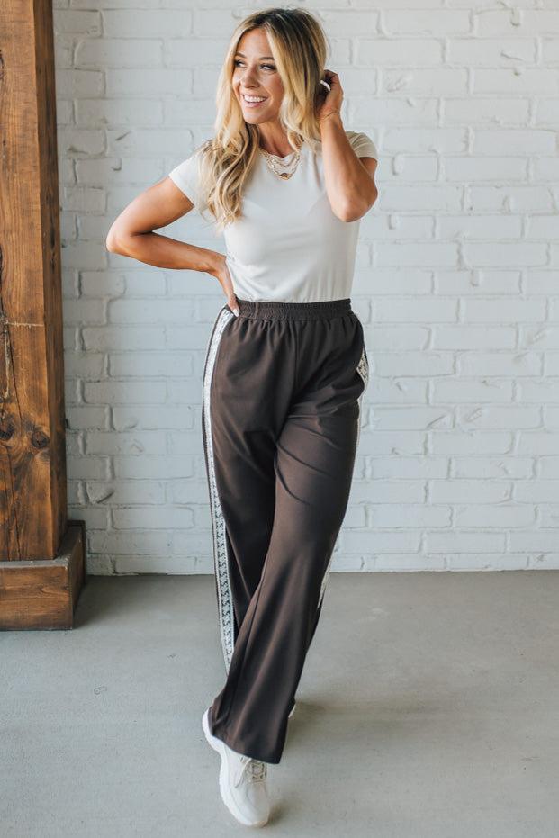 Woman wearing a white top and brown pants with a stripe, standing against a white brick wall.