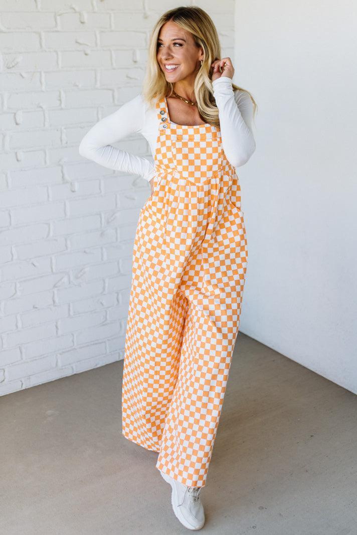 Woman wearing a orange checkered wide leg overalls against a white brick wall.