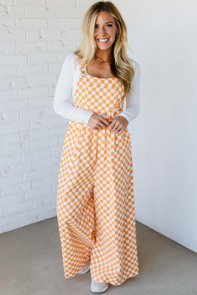 Woman wearing a orange checkered wide leg overalls against a white brick wall.