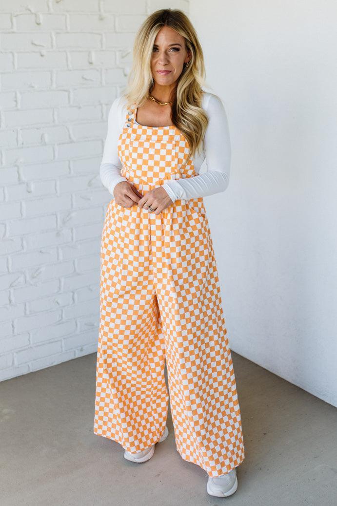 Woman wearing a orange checkered wide leg overalls against a white brick wall.