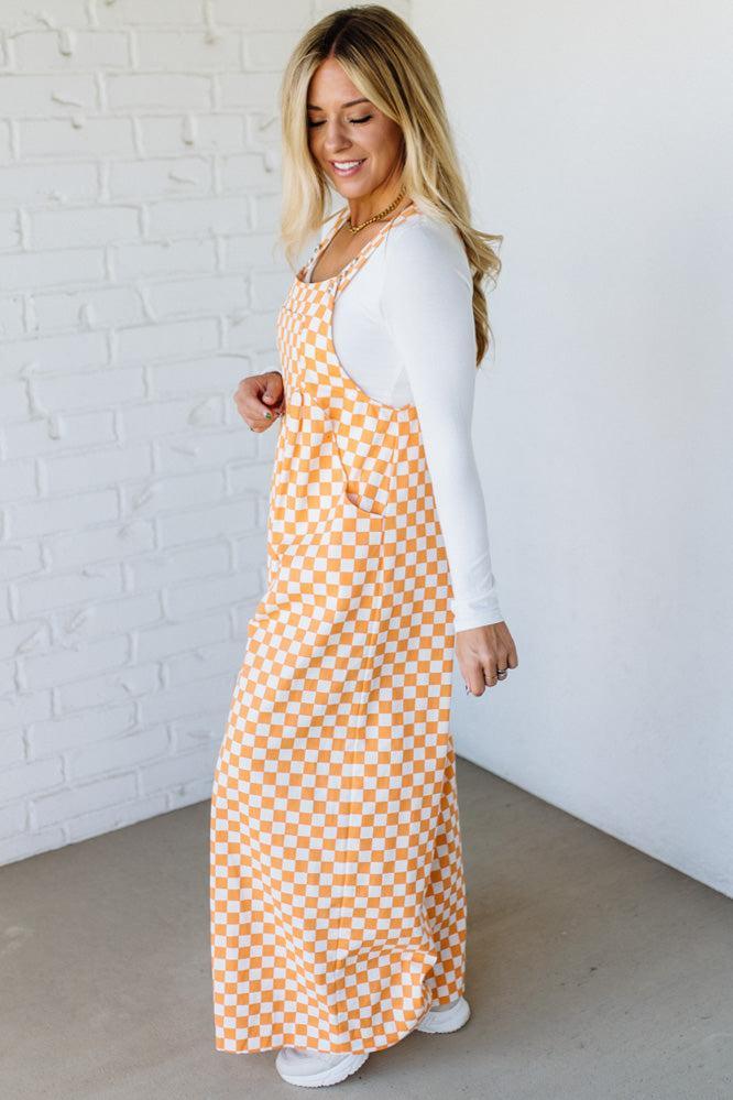 Woman wearing a orange checkered wide leg overalls against a white brick wall.
