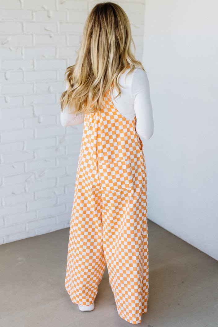 Woman wearing a orange checkered wide leg overalls against a white brick wall.