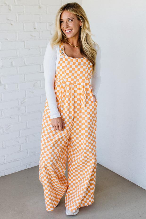 Woman wearing a orange checkered wide leg overalls against a white brick wall.