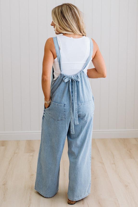 Woman wearing a denim jumpsuit with a white top against a light wooden floor and wall background
