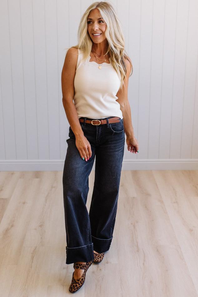 Woman wearing a white tank top and dark blue jeans standing in a room with a light wooden floor and white walls.