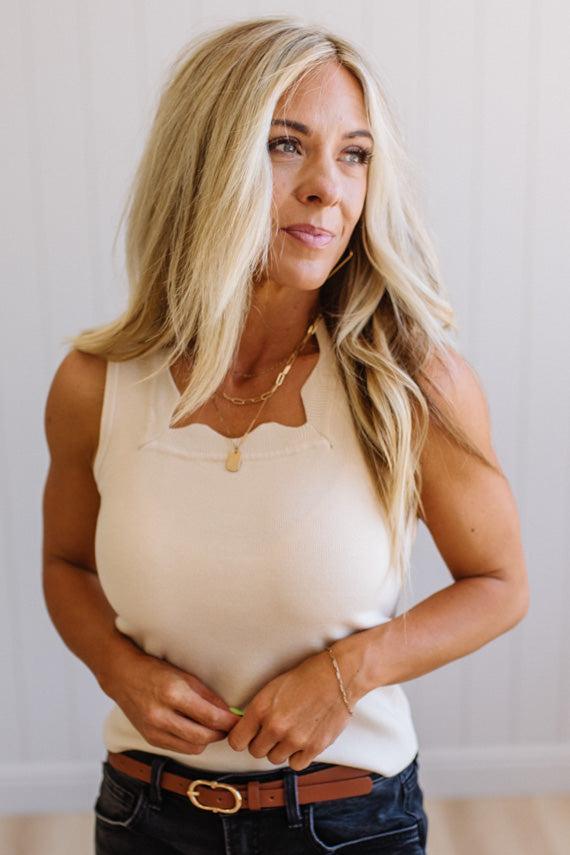 Woman wearing a beige sleeveless top and dark jeans against a plain background