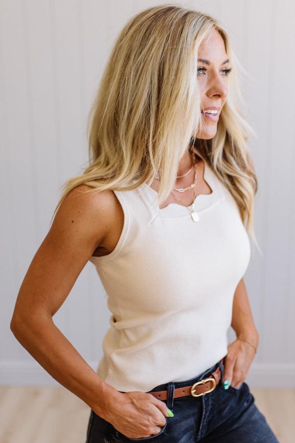 Woman wearing a white sleeveless top and dark jeans against a plain background