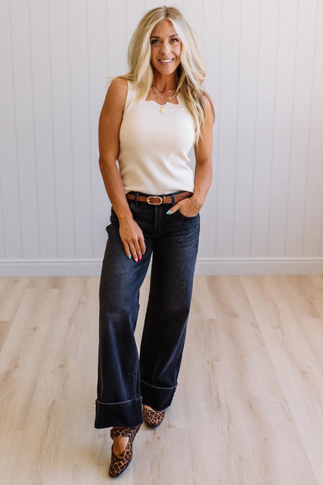 Woman wearing a white tank top and dark blue jeans with a belt, standing in a room with light wooden flooring and a white wall.