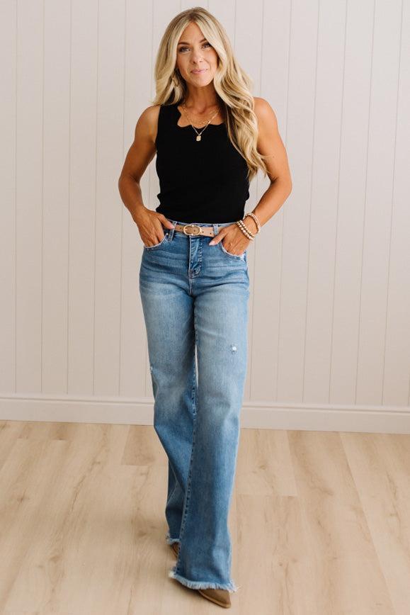 A woman modeling a black sleeveless top and light-wash blue flare jeans with a distressed knee detail, standing in a room with light wood floors and white paneled walls.