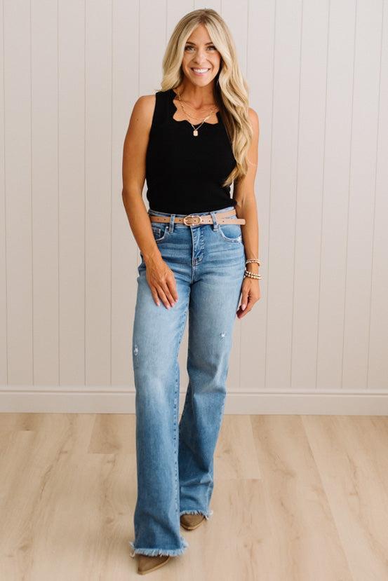 A woman modeling a black sleeveless top and light-wash blue flare jeans with a tan belt, standing against a white paneled wall and light wood floor.