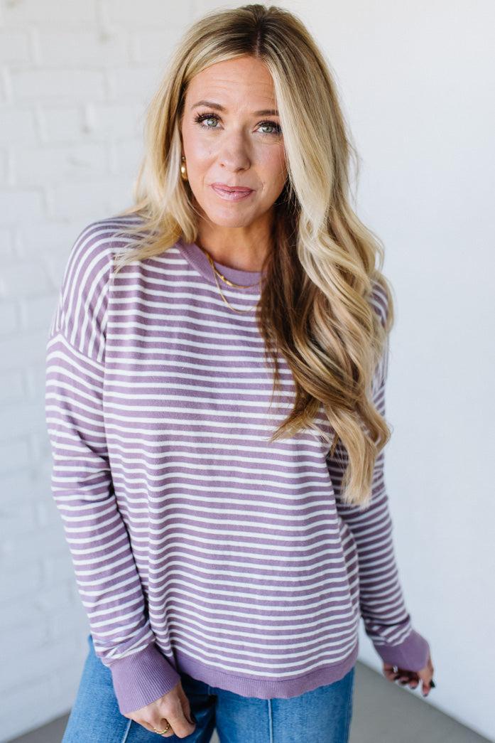 Blonde woman against a white wall wearing a lavender and white striped sweater with blue jeans and sneakers.