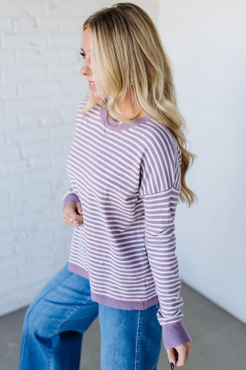 Blonde woman against a white wall wearing a lavender and white striped sweater with blue jeans and sneakers.