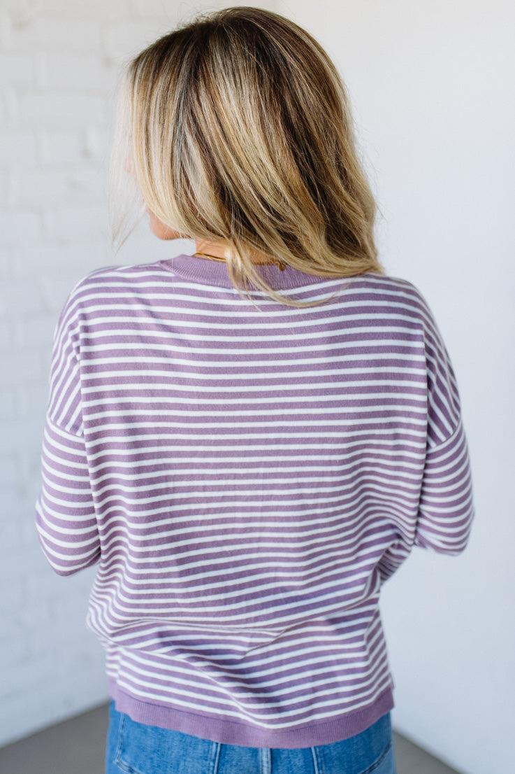 Blonde woman against a white wall wearing a lavender and white striped sweater with blue jeans and sneakers.