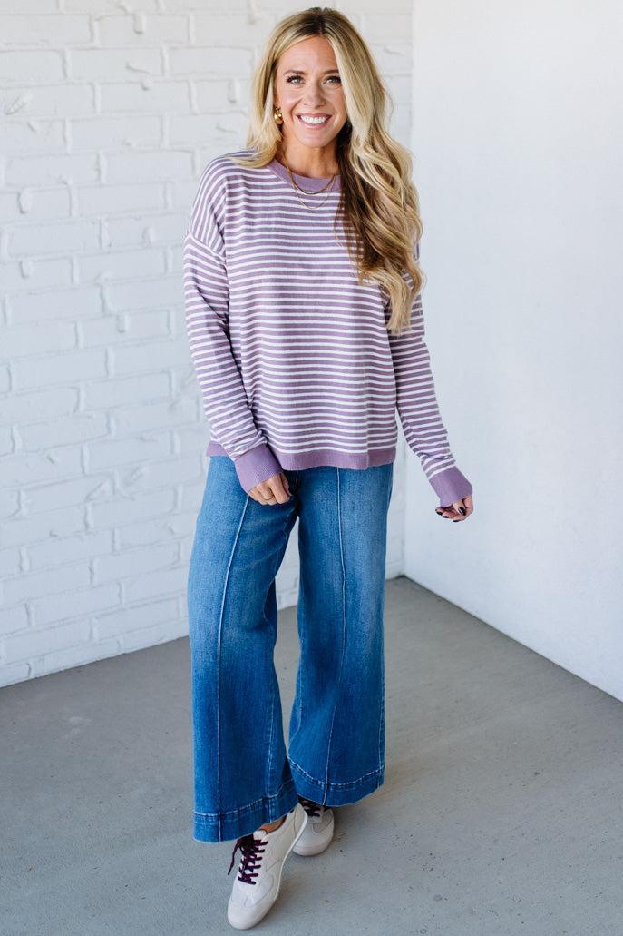 Blonde woman against a white wall wearing a lavender and white striped sweater with blue jeans and sneakers.