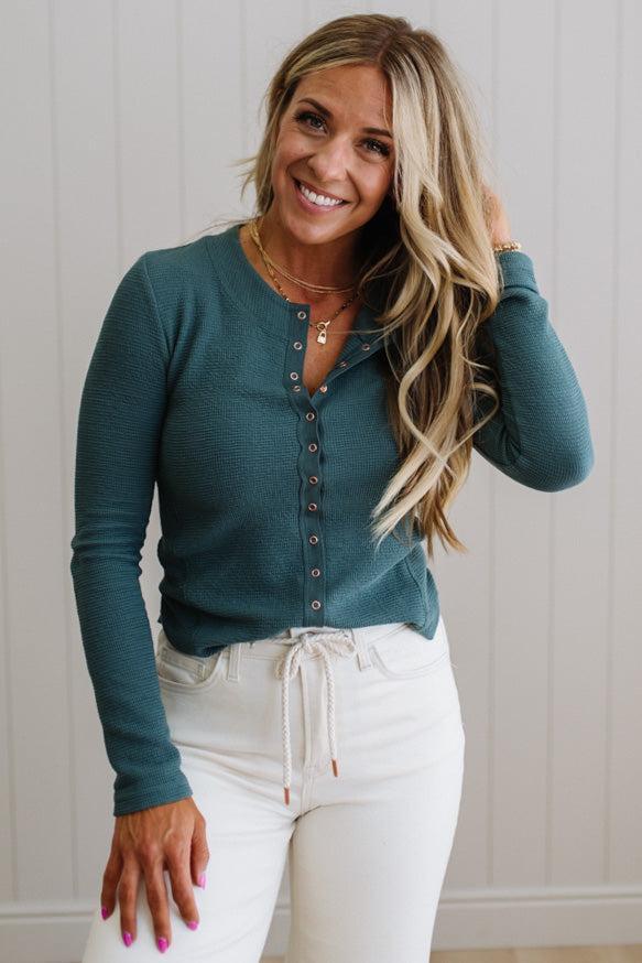 Woman wearing a teal long-sleeve top and white pants against a light background