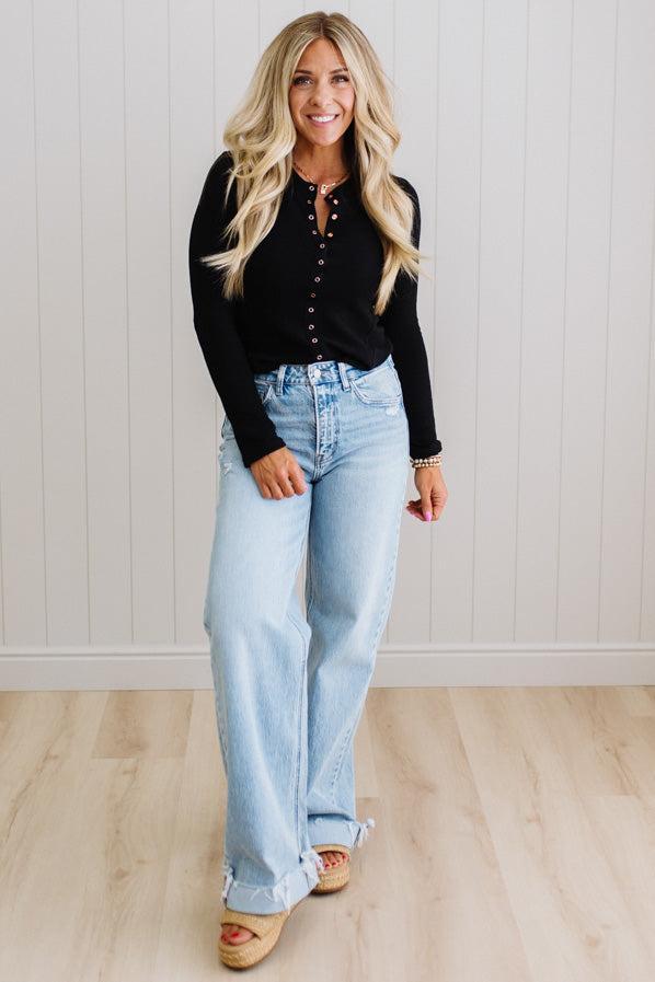 Woman wearing a black long-sleeve top and light blue jeans standing against a white paneled wall.