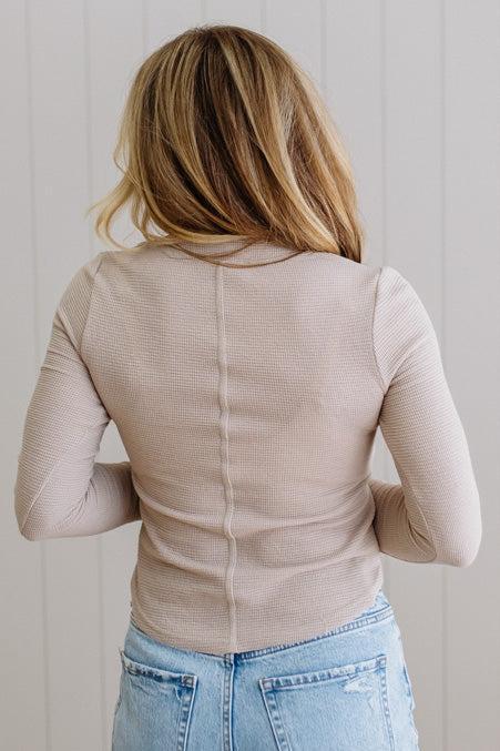 Person wearing a beige ribbed sweater and blue jeans against a light wooden panel background