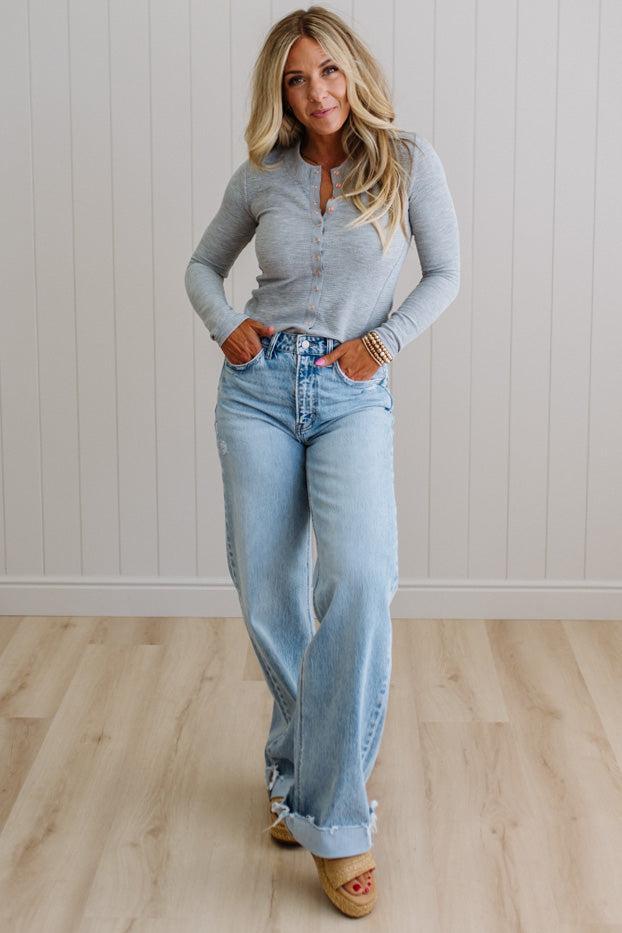 Woman wearing a gray long-sleeve top and light blue jeans standing in a room with wooden flooring and a white paneled wall.