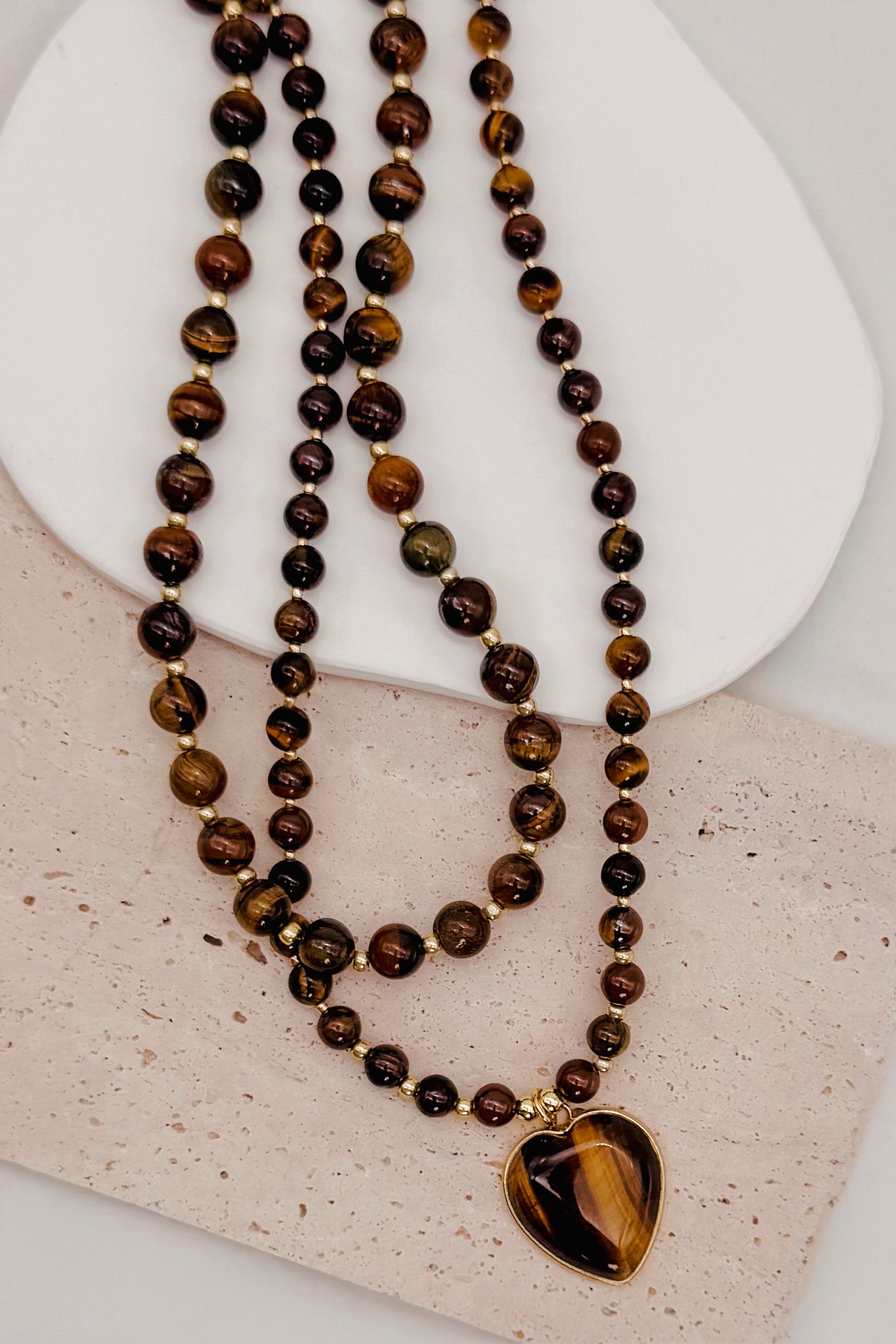 A double-strand beaded necklace featuring tiger's eye beads in shades of brown, black, and gold, with a heart-shaped tiger's eye pendant in a gold-tone bezel, displayed on a textured light beige surface.