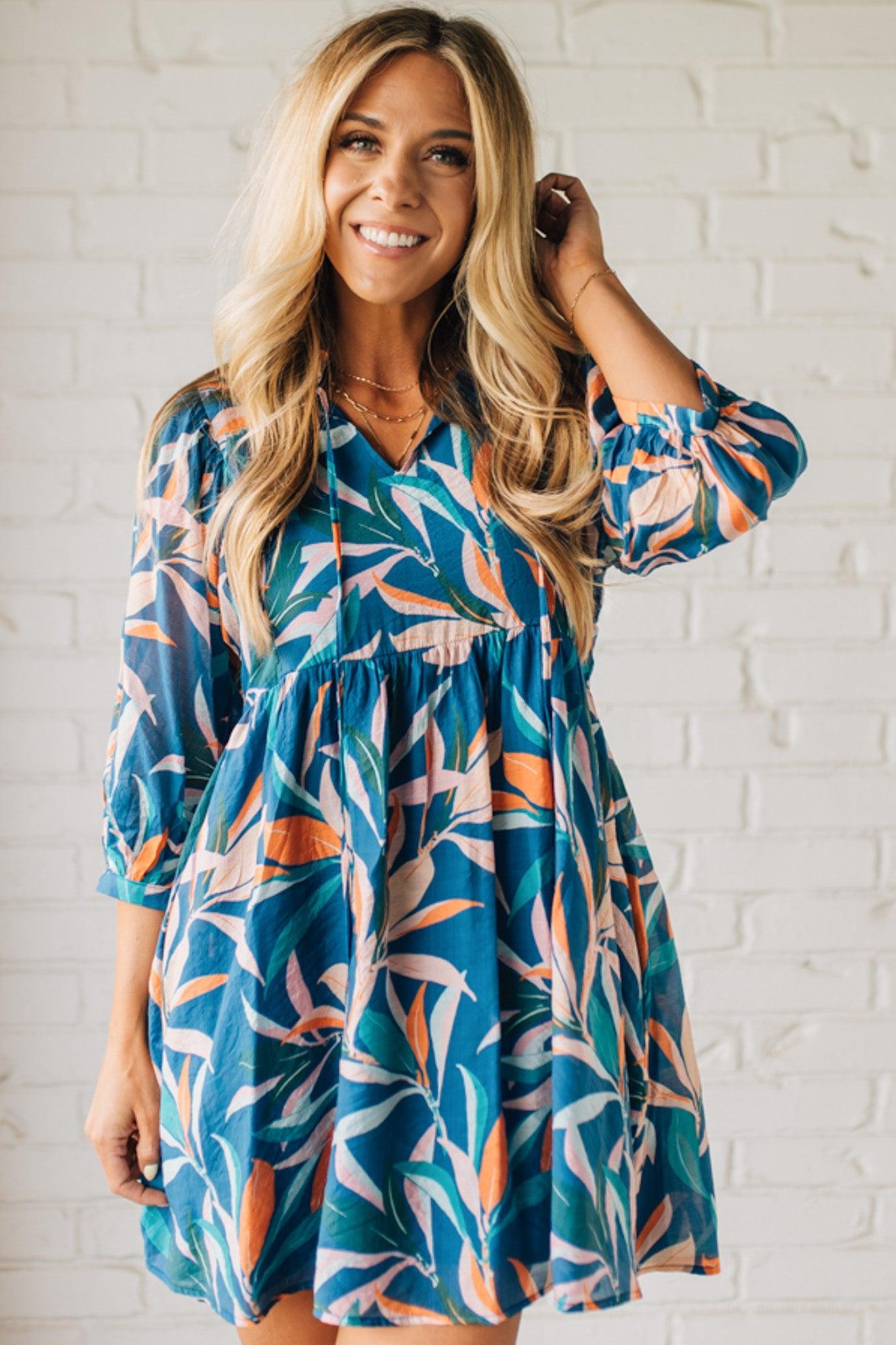 Blonde woman wearing a 3/4 sleeves leaf print dress with front tie neck detail, ruffled neckline, side pockets.