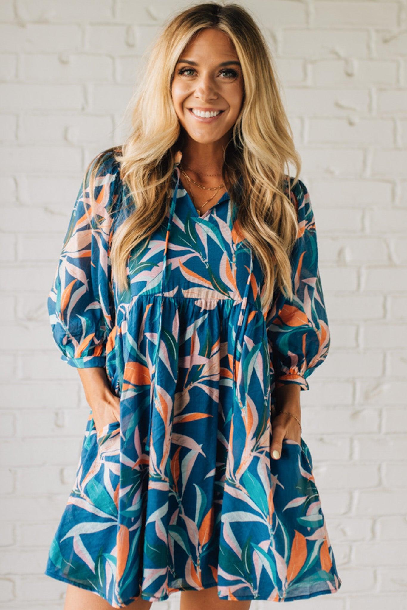 Blonde woman wearing a 3/4 sleeves leaf print dress with front tie neck detail, ruffled neckline, side pockets.