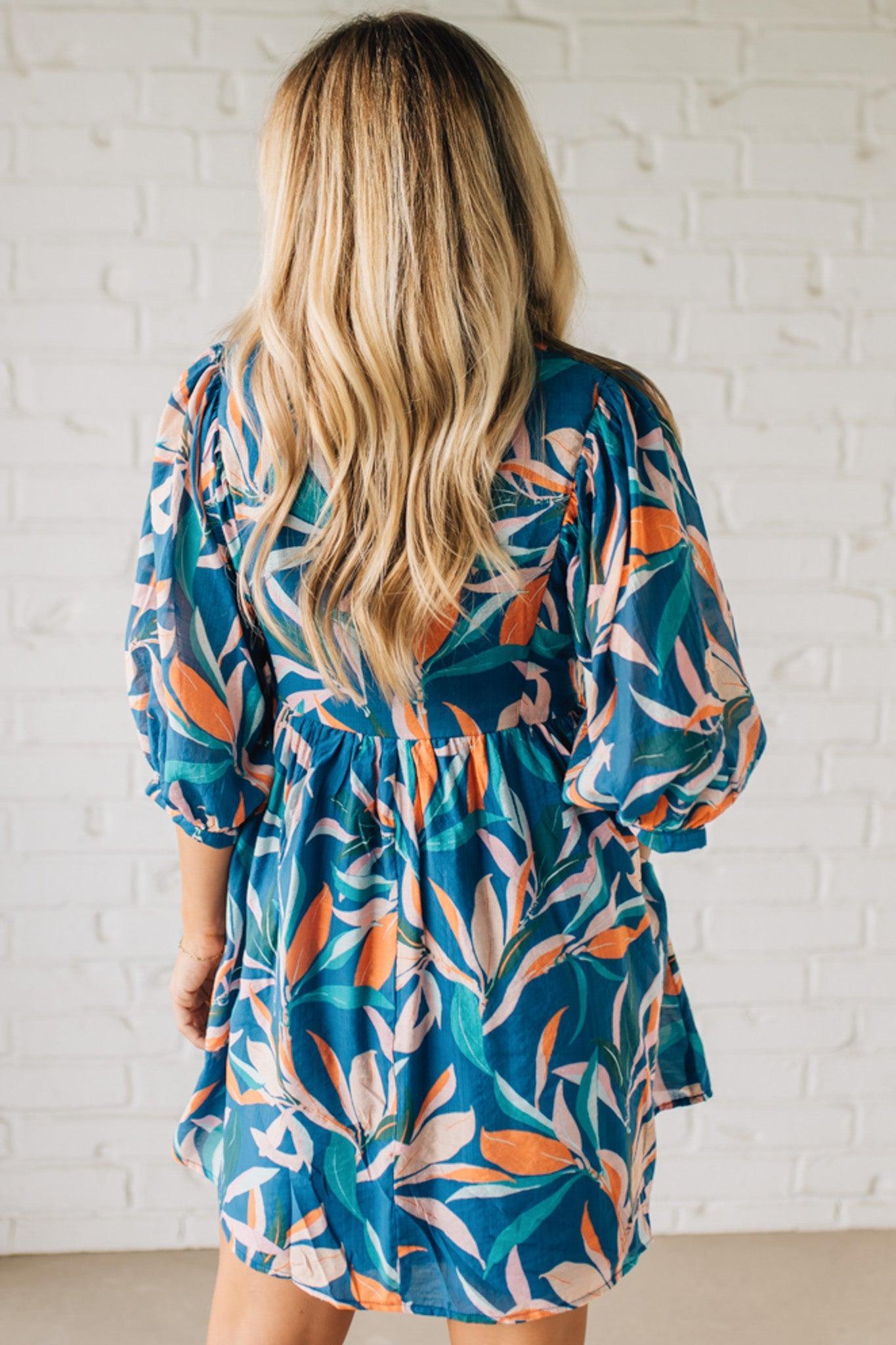 Blonde woman wearing a 3/4 sleeves leaf print dress with front tie neck detail, ruffled neckline, side pockets.