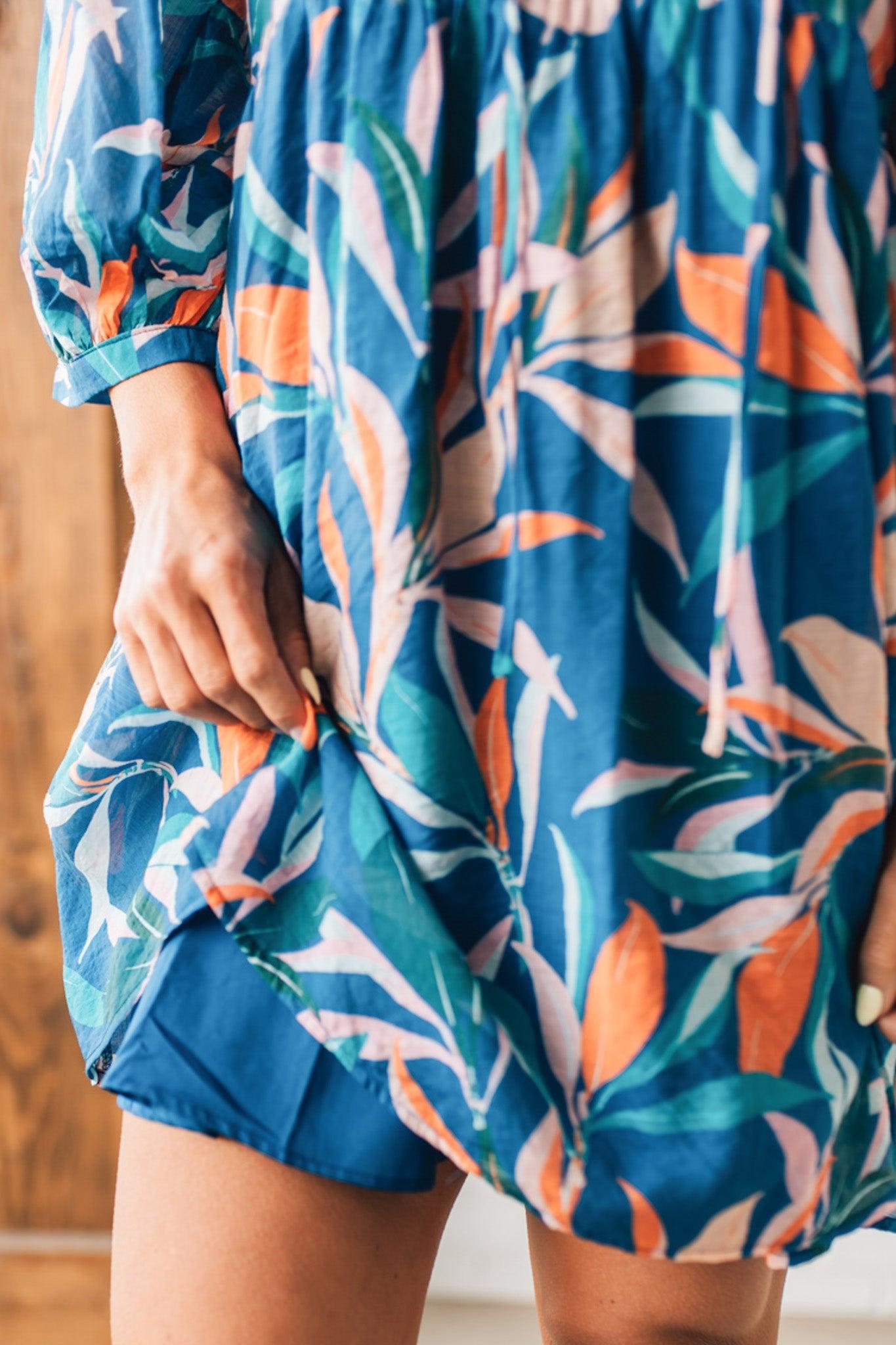 Blonde woman wearing a 3/4 sleeves leaf print dress with front tie neck detail, ruffled neckline, side pockets.