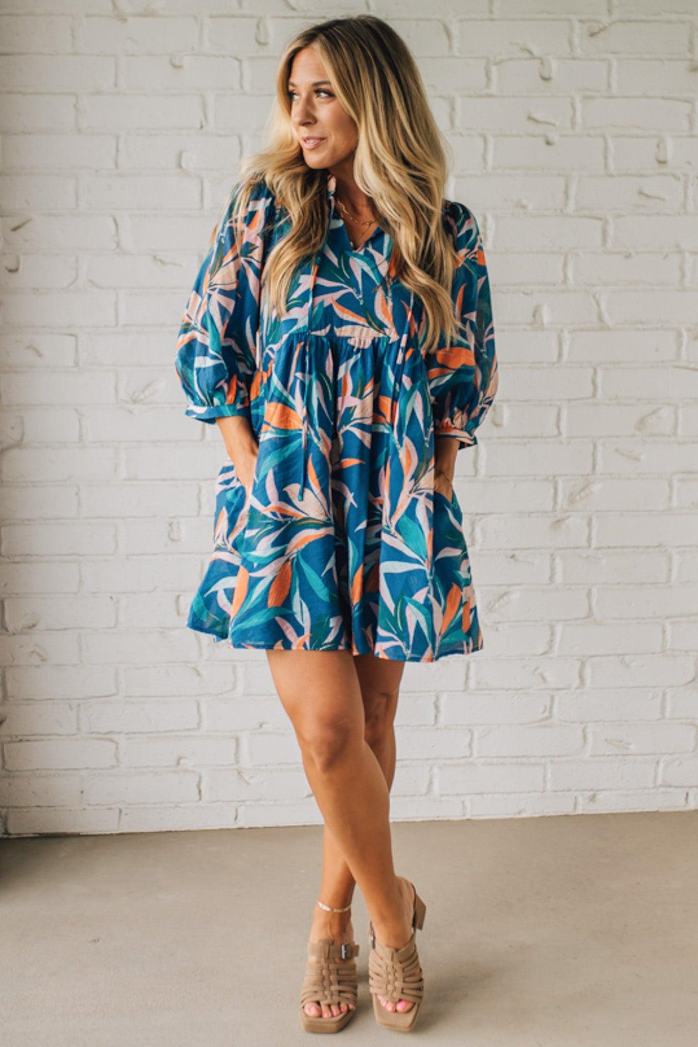 Blonde woman wearing a 3/4 sleeves leaf print dress with front tie neck detail, ruffled neckline, side pockets.