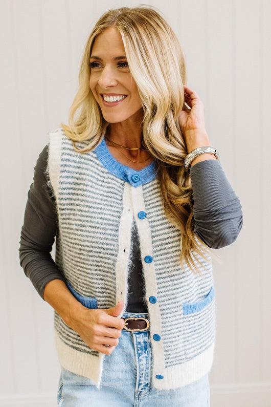 BLONDE WOMAN IN LIGHT JEANS WEARING A DARK GREY TOP WITH A FUZZY SWEATER VEST THAT HAS BLUE TRIM CONTRAST AND BUTTONS, AND A CREAM AND GREY STRIPED BODY.