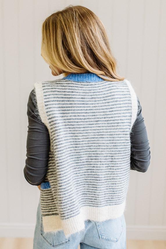 BLONDE WOMAN IN LIGHT JEANS WEARING A DARK GREY TOP WITH A FUZZY SWEATER VEST THAT HAS BLUE TRIM CONTRAST AND BUTTONS, AND A CREAM AND GREY STRIPED BODY.