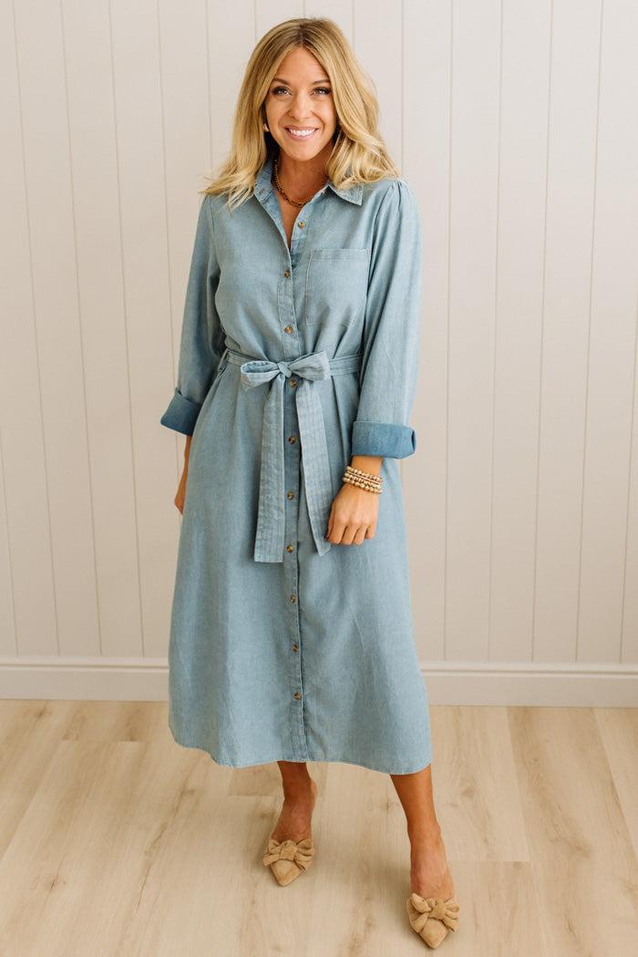 Woman wearing a light blue denim dress standing against a white paneled wall.