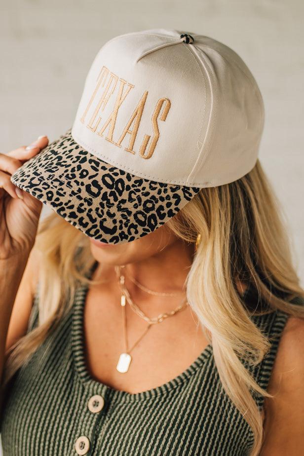 Blonde woman wearing a cream colored trucker hat with leopard print brim, and gold TEXAS embroidered at the front.