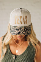 Blonde woman wearing a cream colored trucker hat with leopard print brim, and gold TEXAS embroidered at the front.