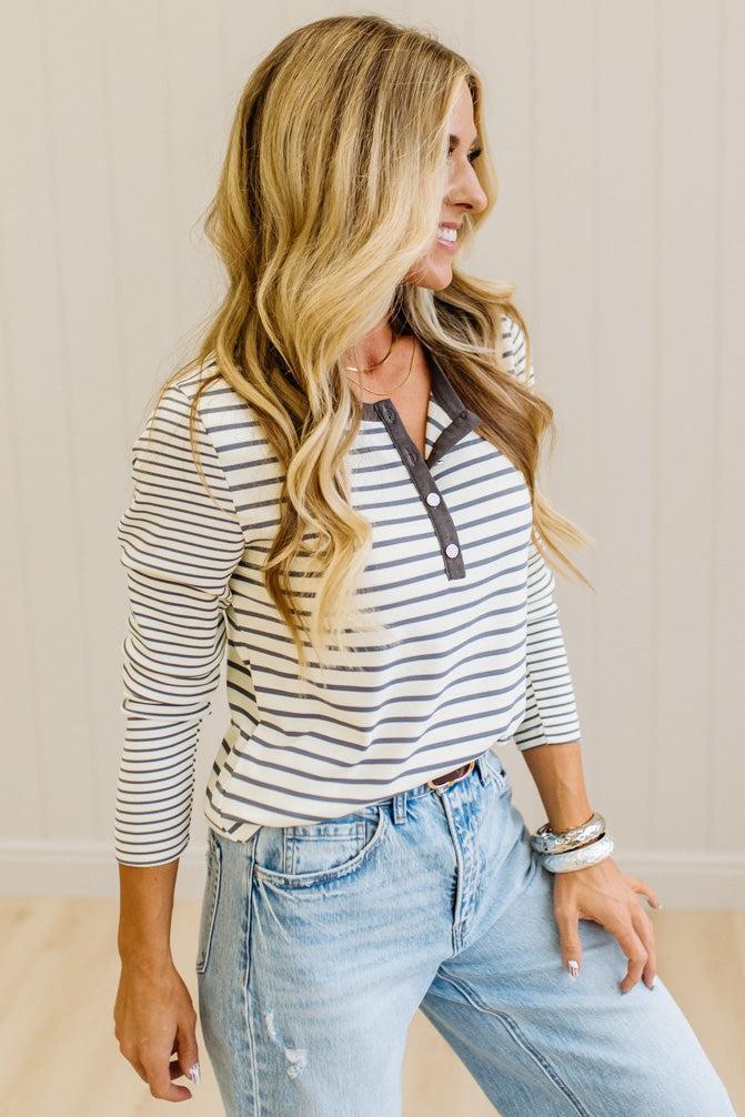 Blonde woman in a brightly lit studio wearing light wide leg jeans, dark clogs, and a striped henley top in cream and charcoal.