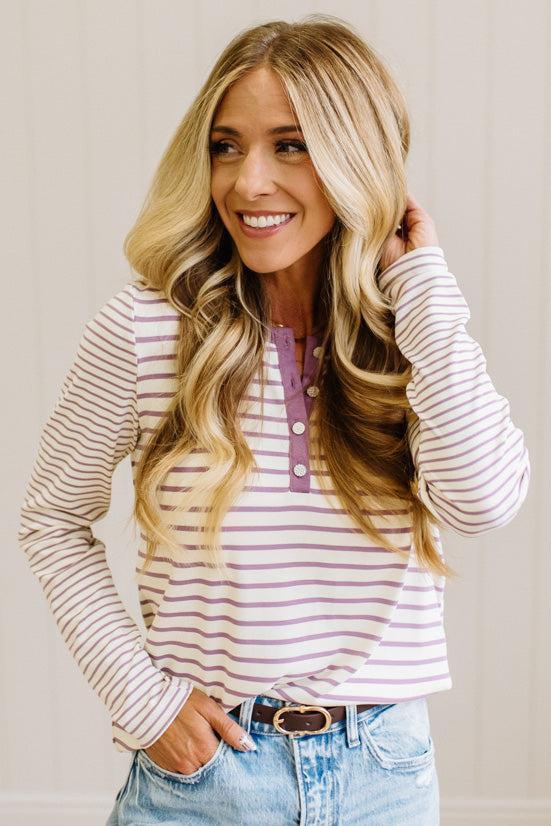 Blonde woman in a brightly lit studio wearing light wide leg jeans, dark clogs, and a striped henley top in cream and lavender.