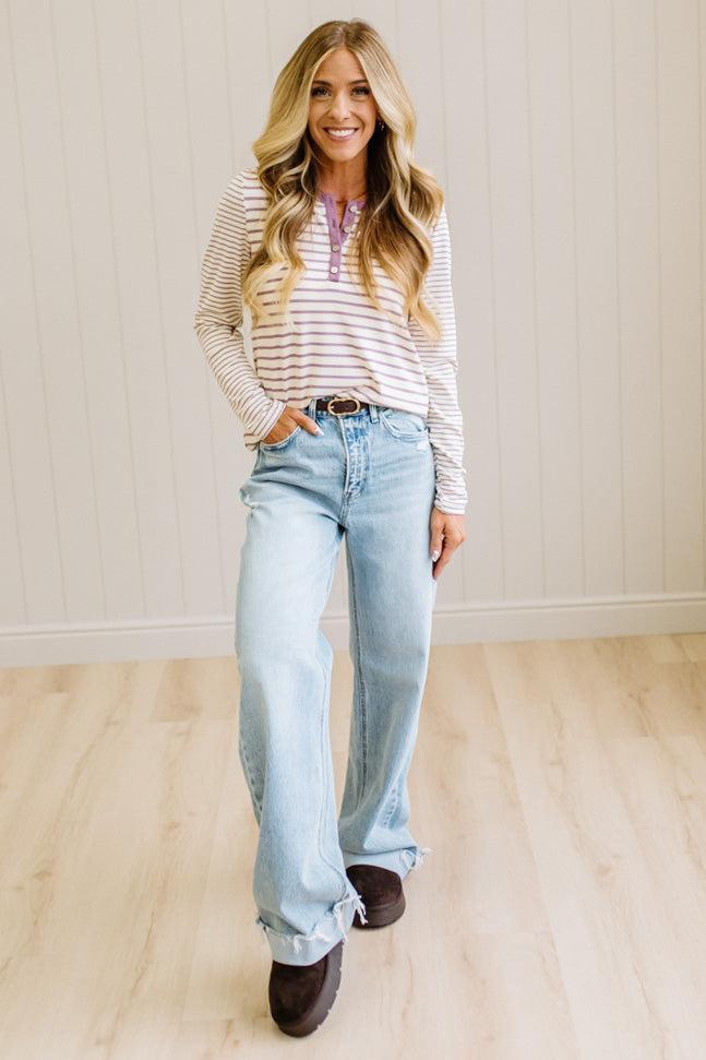 Blonde woman in a brightly lit studio wearing light wide leg jeans, dark clogs, and a striped henley top in cream and lavender.