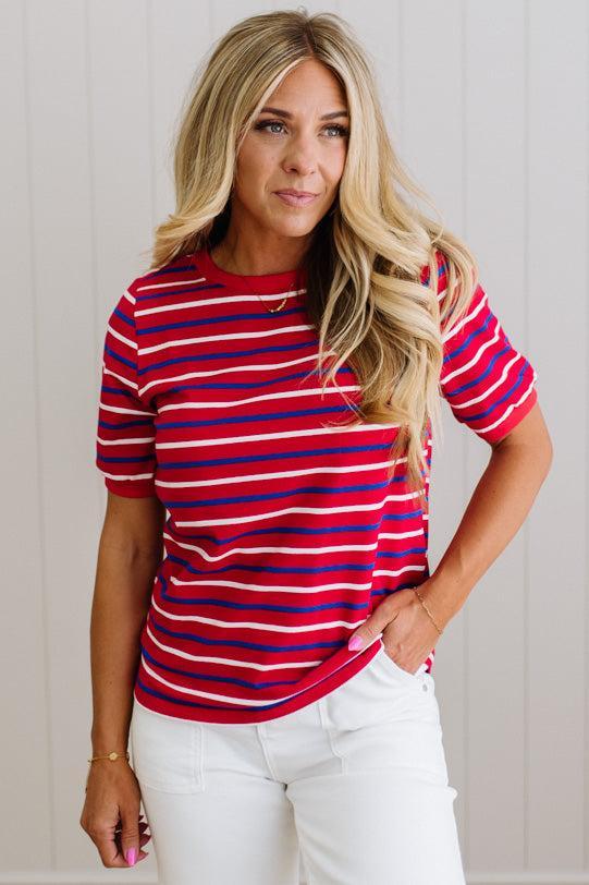 Woman wearing a red and blue striped shirt with white pants against a light wooden panel background