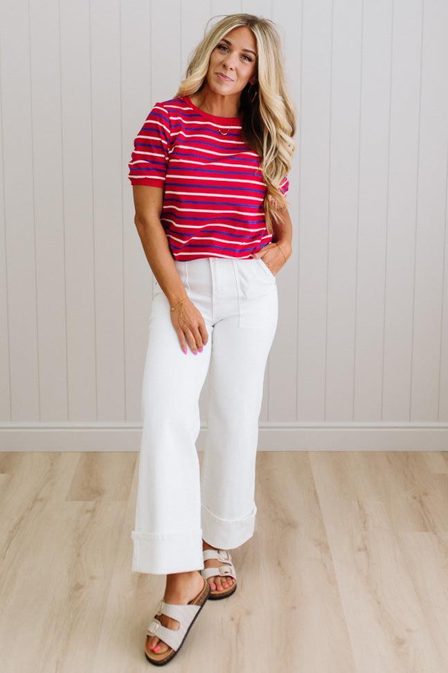 Woman wearing a red and blue striped shirt and white pants standing against a light wooden paneled wall.