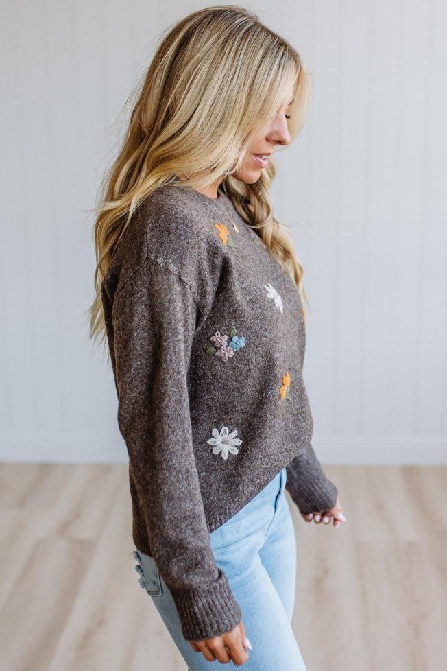 A woman wearing a brown knit sweater with colorful embroidered flowers and white daisy details, paired with light blue jeans, standing in a bright room with light wood floors and white paneled walls.