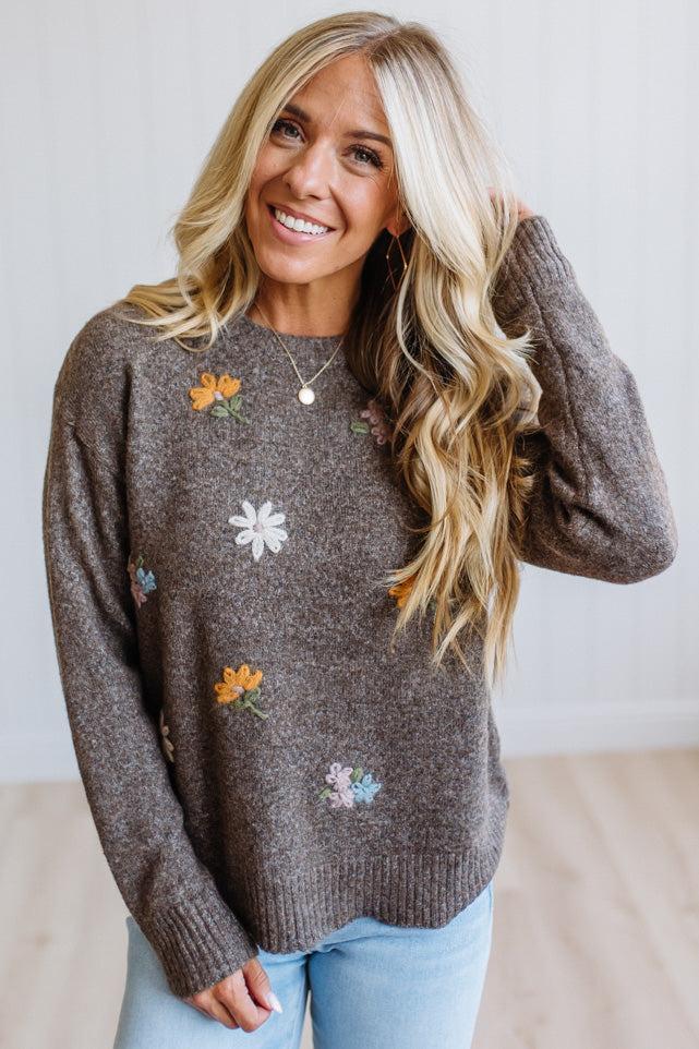 A smiling blonde woman wearing a brown knit sweater with colorful embroidered flowers, paired with light blue jeans, standing against a bright white wall.