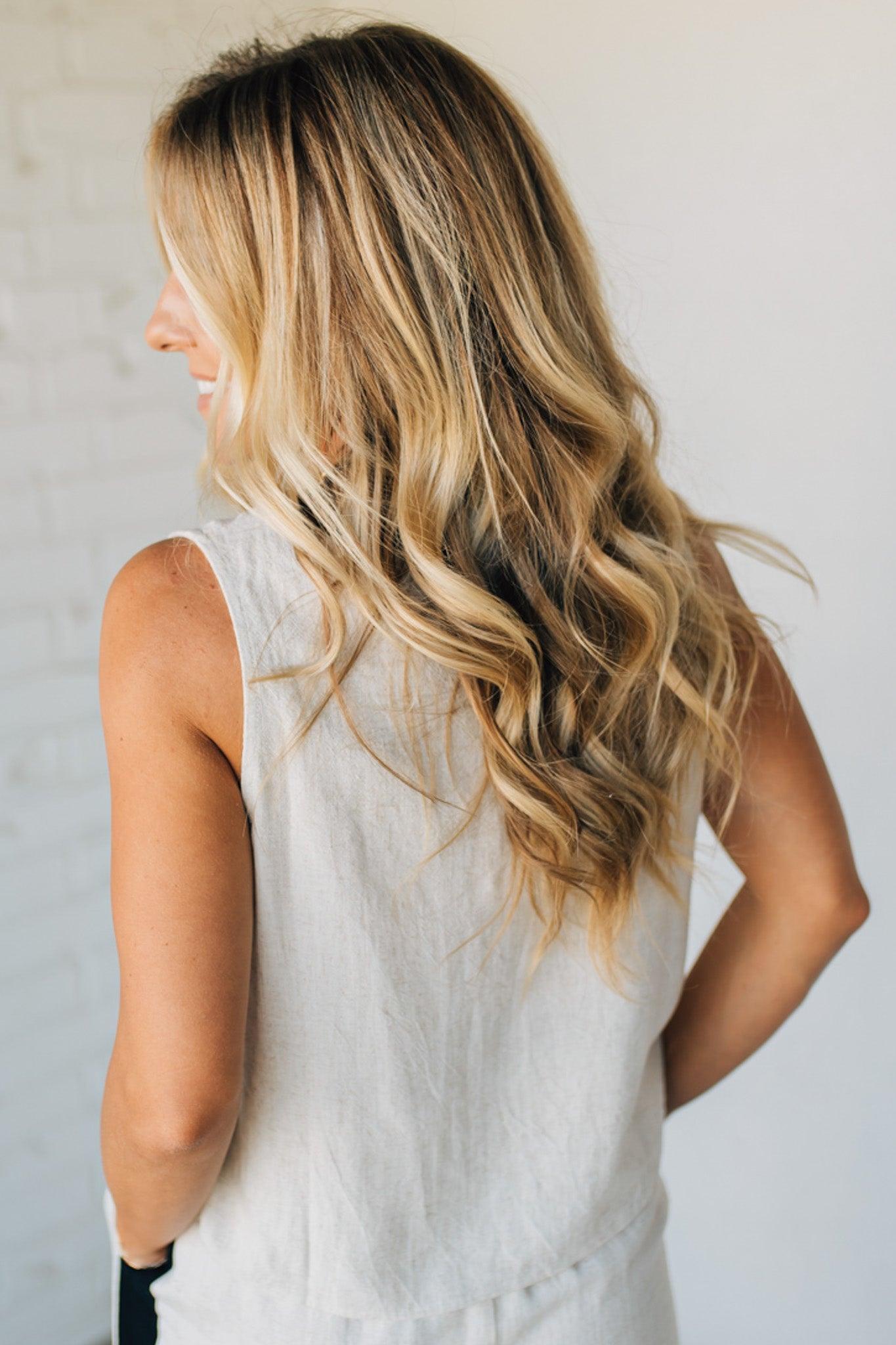 Woman with long, wavy blonde hair wearing a light-colored sleeveless top against a neutral background