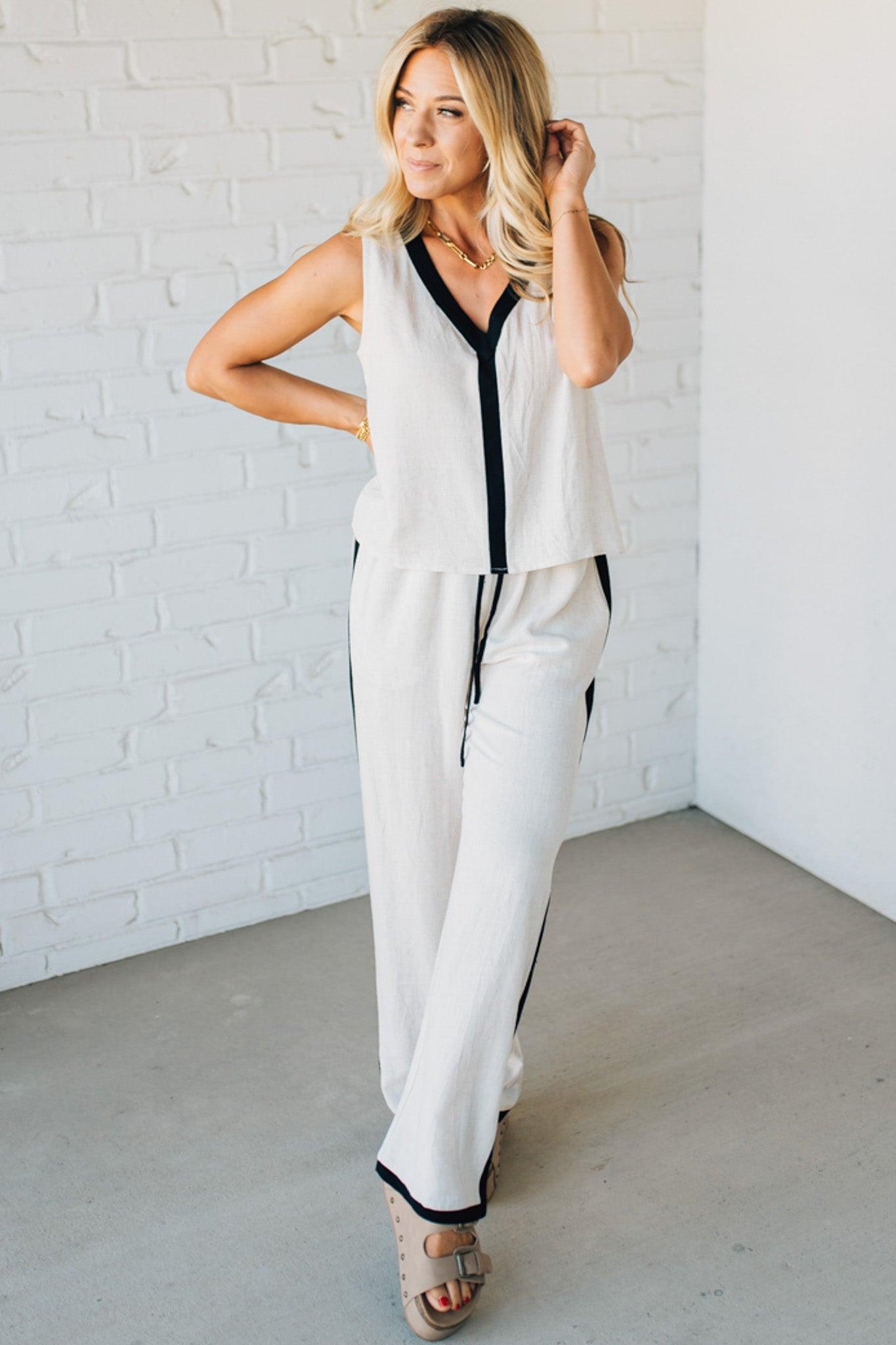 Woman wearing a white sleeveless top and pants with black trim against a white brick wall.