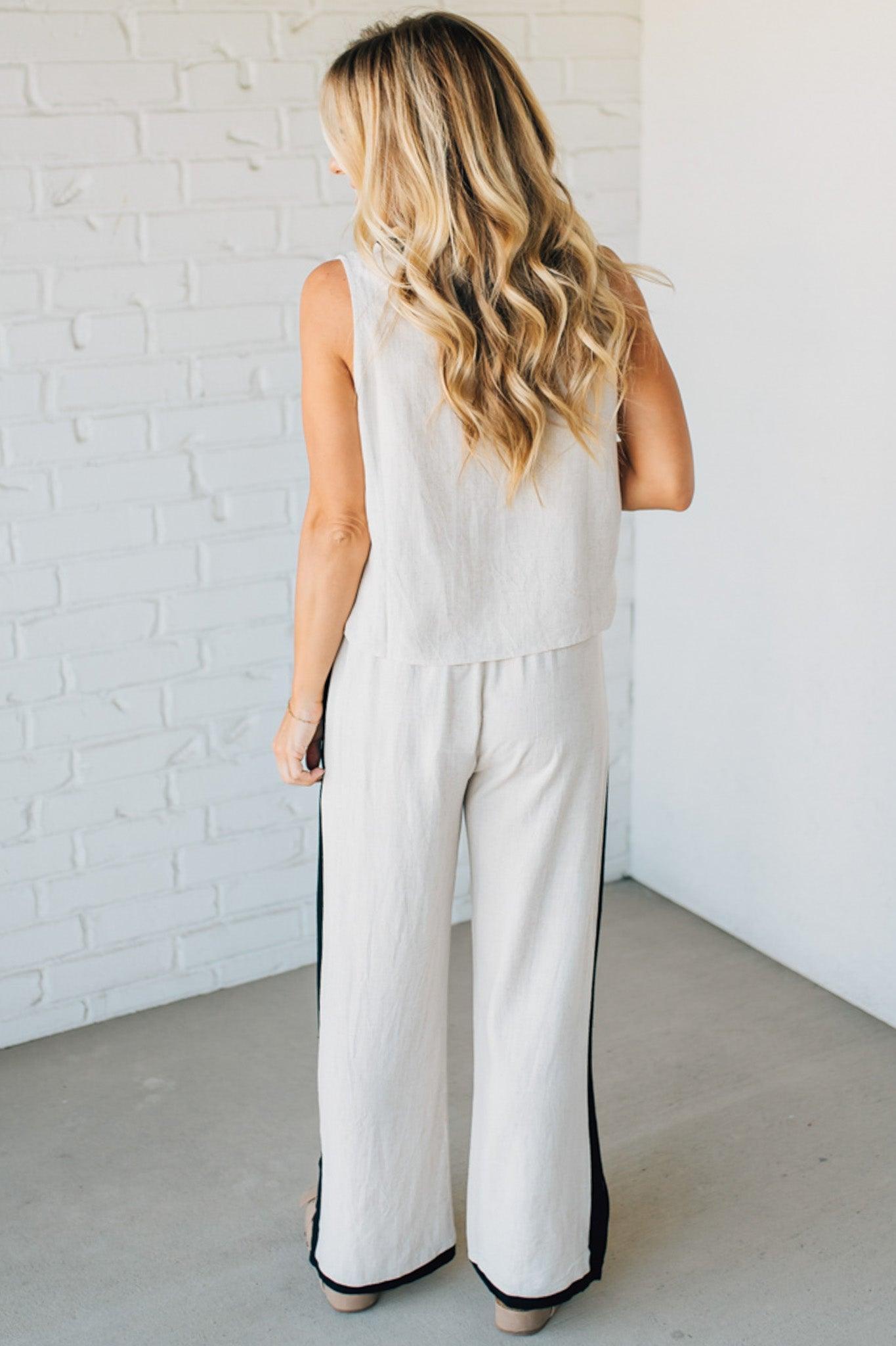Woman wearing a white sleeveless top and pants with black accents against a white brick wall.