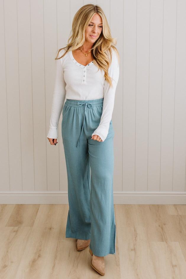 Woman wearing a white long-sleeve top and light blue wide-leg pants against a beige wall.